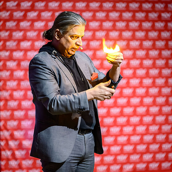 Mann auf Bühne. Über seiner linken Hand ist eine Flamme.
