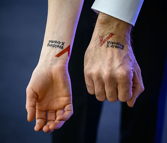 Willkommen beim Tag der Schweiß- und Prüftechnik! Das Motto: Welding X-treme im gediegenen Rahmen:<br /> Zwei tätowierte Arme in Nahaufnahme. Es ist daselbe Tattoo mit der Aufschrift: Welding X-treme.