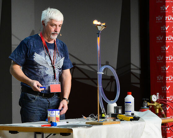 Zum Ende wurde es noch einmal feurig! Martin Kojan (Arbeitssicherheit & Brandschutz) mit dem Vortrag Sicher Arbeiten in Ex-Bereichen.<br /> Mann führt auf Bühne ein Experiment mit Feuer durch