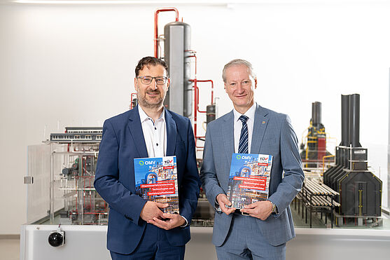 2 Männer haben das neue Buch Chemieverfahrenstechnik in der Hand und stehen vor einer Miniaturausgabe, einem Modell der OMV Raffinerie Schwechat