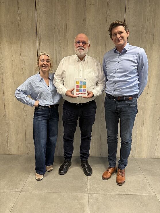 v. l. n. r.: Annelore Vanhoof (Marketing TÜV AUSTRIA BELGIUM), Philippe Dewolfs (Technology Manager OKcert TÜV AUSTRIA BELGIUM), Kamiel Vanderlinden (General Manager TÜV AUSTRIA BELGIUM) <br /> Eine Frau und 2 Männer stehen vor einer Wand. In der Mitte steht der Buchautor mit einer Ausgabe des neuen Buches.