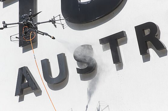 Drohne reinigt TÜV AUSTRIA Logo mit einem Wasserstrahl.