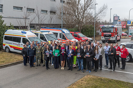 Gruppenbild vor einem Gebäude mit Einsatzfahrzeugen im Hintergrund 