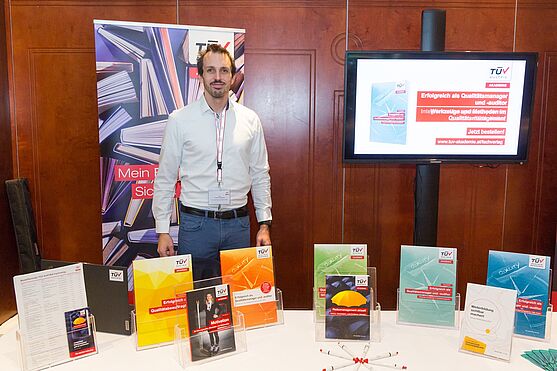 Auch der TÜV AUSTRIA Fachverlag war gutgelaunt mit einem Ausstellerstand vor Ort vertreten!<br /> Lächelnder Herr im Hemd hinter einem Ausstellerstand.