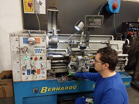 Vor einer Drehmaschine zeigt ein Mann in blauem Pulli mit Schutzbrille und Handschuhen einen Safety-Check vor