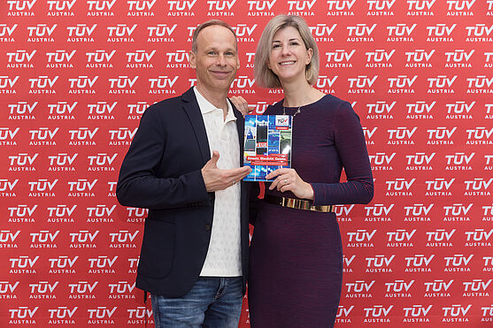 Ein Mann und eine Frau mit Buch vor einem Fotohintergrund mit Logos
