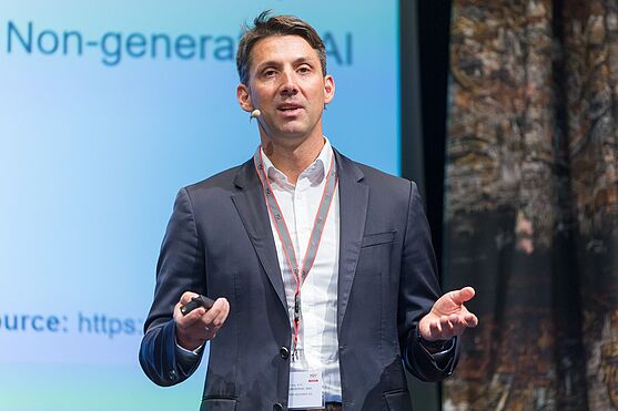 Als nächstes sprach Martin Hofstädtner, TÜV AUSTRIA über AI-Anwendungen zur Effizienzsteigerung von Qualitätsmanagementprozessen.<br /> Herr auf der Bühne hält einen Vortrag. Hinter ihm die Präsentation auf einer Leinwand.