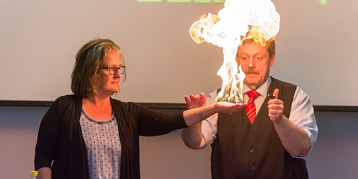 Physiker Werner Gruber mit einer Freiwilligen aus dem Publikum. Über ihrer flach ausgestreckten Hand, ist eine Flamme.