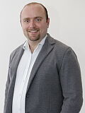 Stefan Grüneis, ein Mann in formeller Kleidung, steht vor einer weißen Wand und vermittelt einen professionellen Eindruck.