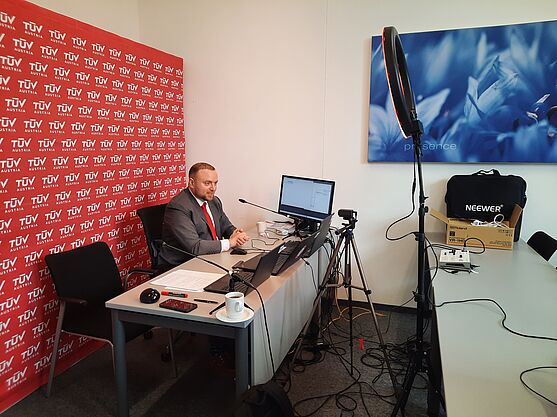 Herr im Anzug sitzt an einem Schreibtisch. Hinter ihm eine Pressewand, vor ihm ein Laptop inklusive Livestreaming Setup.