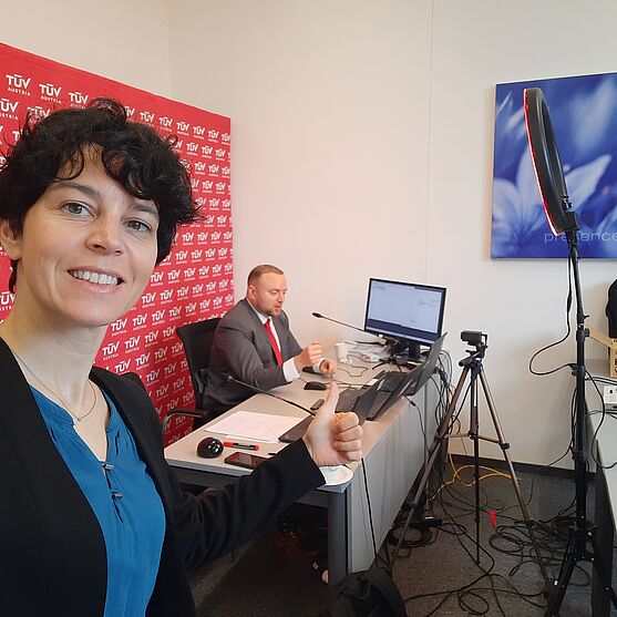 Dame mit erhobenem Daumen. Hinter ihr ein Livestream-Setup und ein Herr im Anzug der vor einem Laptop sitzt.