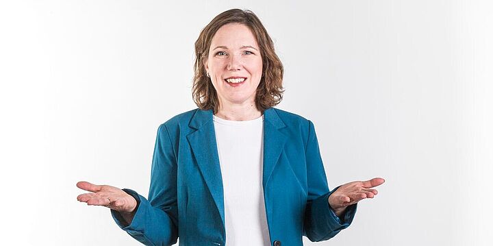 Freundlich lächelnde Dame mit blauem Sakko, die ihre Handflächen nach oben hält.