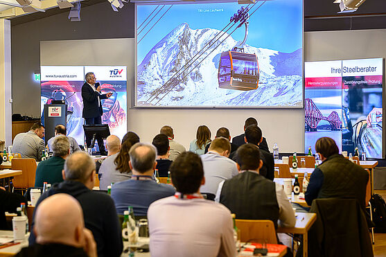 Zurück zum Wesentlichen: Harald Germ (Doppelmayr Seilbahnen GmbH) lieferte spannende Einblicke in Bauteilprüfungen im Seilbahnbau.<br /> Ein Mann im schwarzen Anzug steht vor einer Videowall, auf der eine Seilbahn in den verschneiten Bergen gezeigt wird.