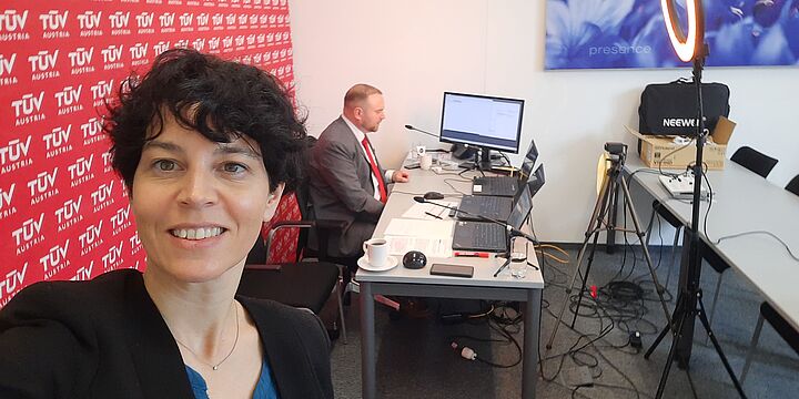 Freundliche lächelnde Dame. Hinter ihr sitzt ein Herr an einem Schreibtisch vor einem Laptop. Sie sind umgeben von Livestreaming-Equipment.