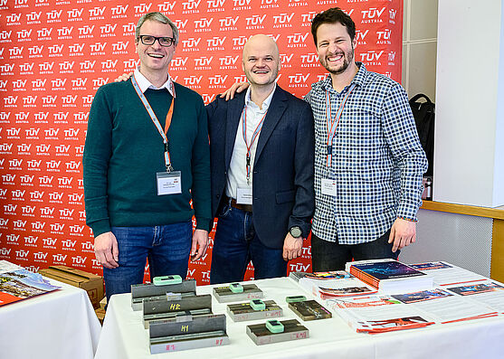 <br /> 3 Männer stehen vor einem Tisch, auf denen Werkstücke und Werbematerial liegt.