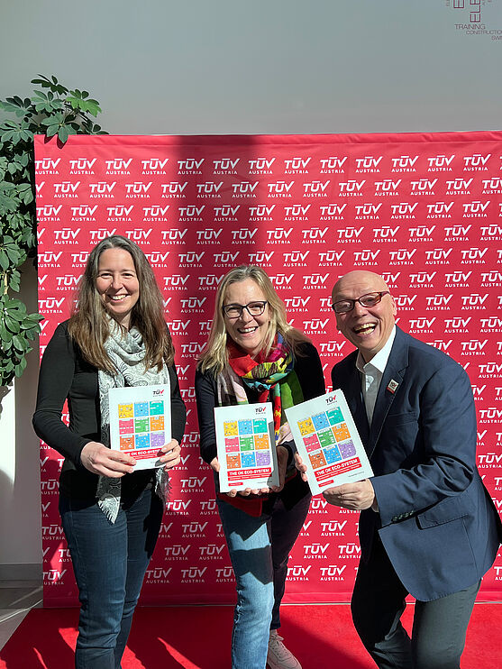 TÜV AUSTRIA Fachverlag freut sich über Abschluss des ersten internationalen Buchprojekts: Judith Martiska (Fachverlagsleitung), Monika Kammerer (Leitung E-Learning & Fachverlag) und Christian Bayer (Geschäftsführer TÜV AUSTRIA Akademie)<br /> Drei Personen, zwei Frauen und ein Mann stehen nebeneinander vor einer roten Wand und halten ein Buch in die Kamera.