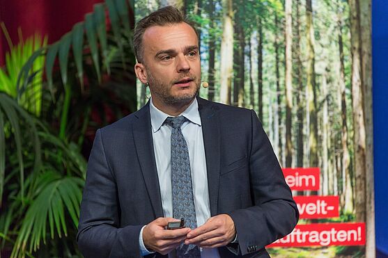 Mann mit Sakko, Hemd und Krawatte hält Vortrag auf der Bühne.