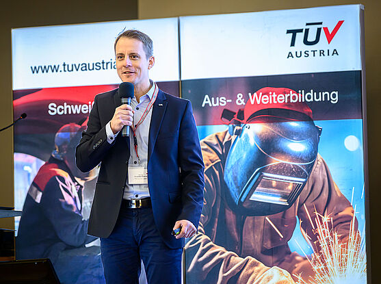Alexander Mastnak (TÜV AUSTRIA GMBH) hielt den ersten Vortrag des Tages: Die neue Bauprodukte-Verordnung: Was Hersteller von Metallbauprodukten jetzt wissen müssen<br /> Ein Mann mit kurzen Haaren und blauem Anzug auf einem Podium. Er spricht in ein Mikro.