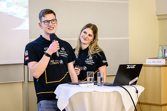 Larissa Röck und Maximilian Kainz (TU Wien Racing) sprachen über 'Formular Student: Engineering on the EDGE' in ihrer Keynote.<br /> Ein Mann mit Brille und eine blonde Frau stehen hinter einem Stehtisch mit Mikros in der Hand. Sie tragen gesponserte T-Shirts mit Markenaufschriften.