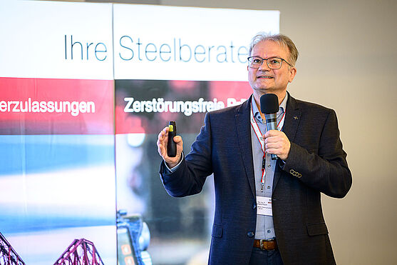 Weiter ging es mit Gerhard Posch (voestalpine böhler welding GmbH) welcher wertvolle Informationen zum Themenfeld Health and Safety in der Schweißtechnik lieferte.<br /> Ein Mann mit Brille und dunklem Anzug erklärt auf dem Podium etwas.