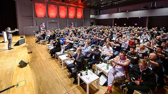 Mann auf Bühne streckt beide Arme aus. Publikum macht es ihm nach.