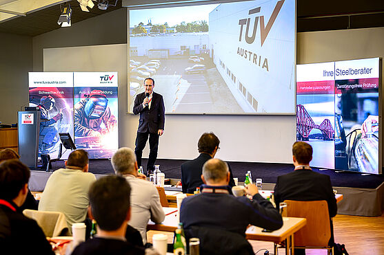 Peter Weinzettl (TÜV AUSTRIA) startete in den Nachmittag. Er moderierte eine 'Liveschaltung' nach Wien ins Prüfzentrum.<br /> Ein Mann mit Mikro steht auf der Bühne. Hinter ihm läuft ein Video auf einer Leinwand. Es zeigt ein Gebäude mit der Aufschrift 'TÜV AUSTRIA'.