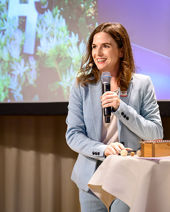 Herzlich willkommen zum TÜV AUSTRIA Wasserstoff-Symposium! Sabine Redlich (TÜV AUSTRIA Akademie) sorgte mit einleitenden Worten für einen freundlichen Empfang.<br /> Dame auf Bühne spricht lächelnd in Handmikrofon