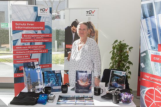 Freundlich lächelnde Dame hinter Messestand der TÜV Trust IT.
