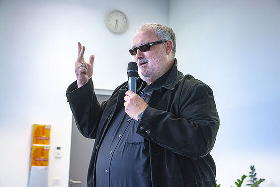 <br /> Mann in schwarz mit Sonnenbrille spricht in Handmikrofon.