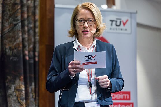 Vortragende mit Brille und Notizen in der Hand, steht auf einer Bühne.