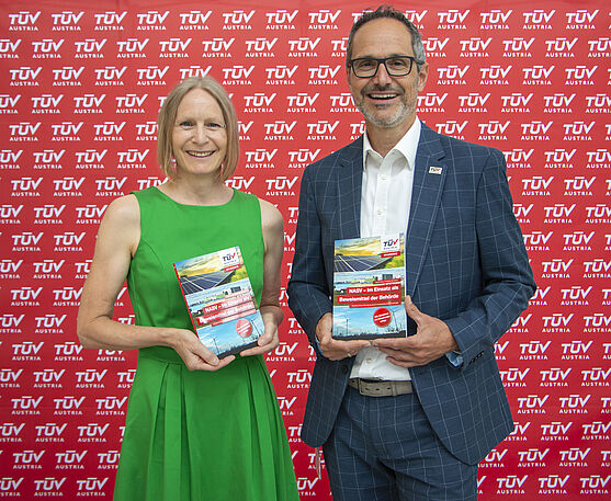 Autorin Ingrid Heinz (im grünen Kleid) und Autor Andreas Schnitzer (im Anzug) stehen mit dem neuen Buch NASV vor einer roten Pressewand