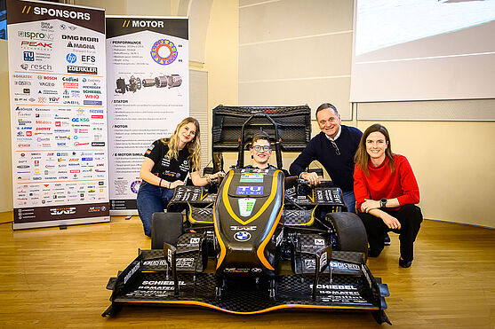Das passende Fahrzeug war DER Hingucker und Fotopunkt des Abends! <br /> Ein schwarzes Rennauto mit gelben Streifen der TU-Wien. Ein Mann mit Brille sitzt im Auto, zwei Frauen und ein Mann knien daneben und lächeln in die Kamera.