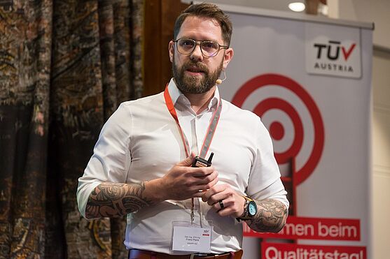 Franz Plank, KNAPP AG mit dem Vortrag: QMS Development Planning: The "Governance Model as an anchor" - How strategy becomes reality!<br /> Vortragender auf der Bühne mit Bart, Brille, aufgestrecktem Hemd und Tattoos an den Unterarmen.