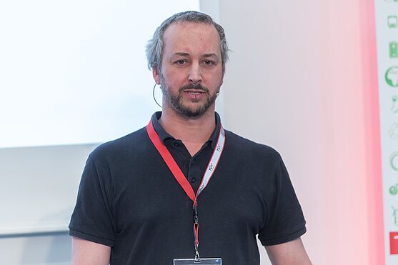 Mann in schwarzem Poloshirt hält Vortrag.