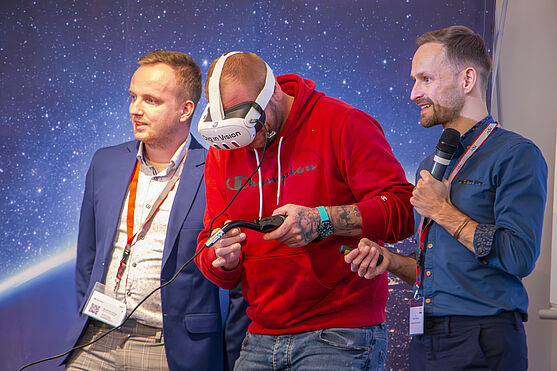 Ausprobieren erwünscht!<br /> Drei Männer auf der Bühner. Einer verwendet ein VR Headset.