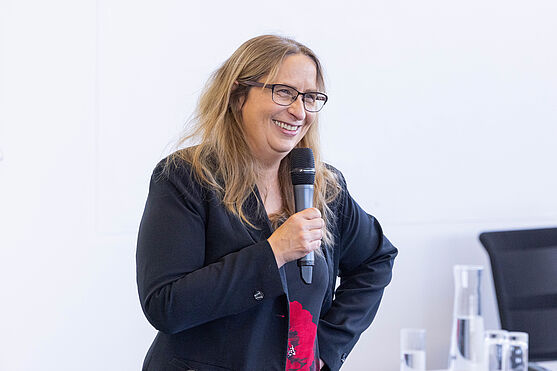 Karin Newald (TÜV AUSTRIA Akademie) hieß die Teilnehmenden herzlich willkommen zum diesjährigen TÜV AUSTRIA Tag der Barrierefreiheitsbeauftragten.<br /> Dame lächelt und hält Handmikrofon