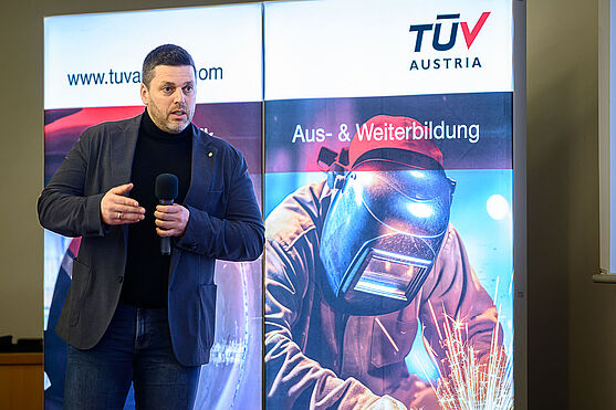 Daniel Irkuf (Kremsmüller Anlagenbau GmbH) im Austausch mit Jochen Bognar (TÜV AUSTRIA GmbH) über den Entwurf der neuen EN ISO 9606: Hersteller im Austausch mit der Zertifizierungsstelle.<br /> Ein Mann im dunklen Anzug und mit kurzen Haaren mitten im Vortrag.