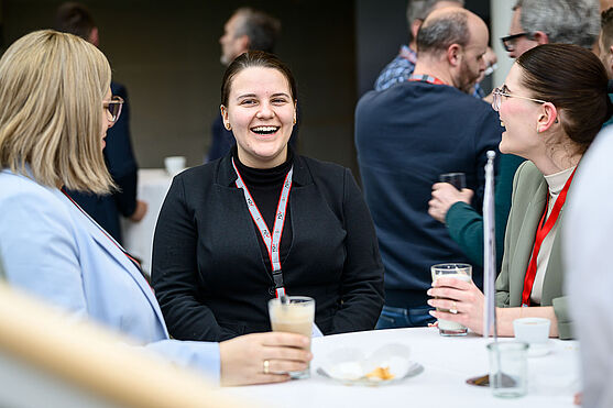 ...munteren Austausch unter Kolleg:innen...<br /> 3 Frauen im Businessoutfit stehen um einen Stehtisch herum und lachen.