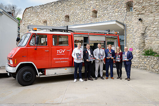 Sechs Männer stehen vor Feuerwehrauto und halten Brandschutzbuch in den Händen.