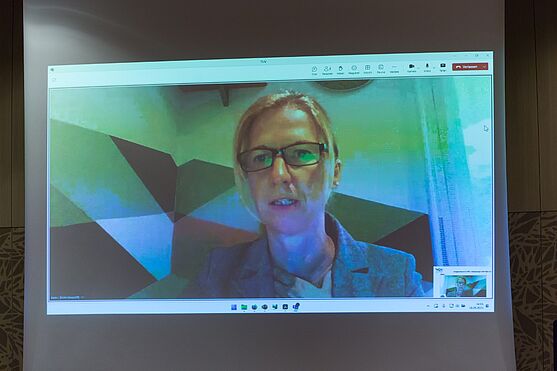 Live zugeschalten war Karin Lenhard von der ERSTE Bank, die über ESG-Projekte in ihrem Unternehmen sprach.<br /> Eine Frau ist per Videocall auf der Leinwand des Vortragssaals zu sehen. Live-online sprach die Referentin Karin Lenhard über ESG-Projekte in der ERSTE Bank.