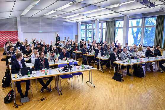 Auf die Plätze...fertig...los geht's mit dem ersten Vortrag!<br /> Ein mit Menschen gefüllter Vortragssaal. Einige der Teilnehmer:innen haben die Hand gehoben.