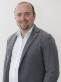 Stefan Grüneis, ein Mann in formeller Kleidung, steht vor einer weißen Wand und vermittelt einen professionellen Eindruck.